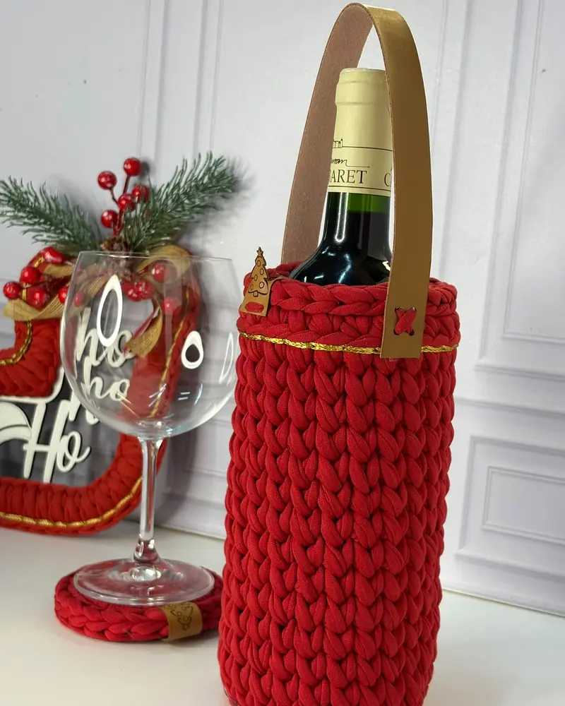 Porta vinho de crochê vermelho com alça de couro, ao lado de uma taça